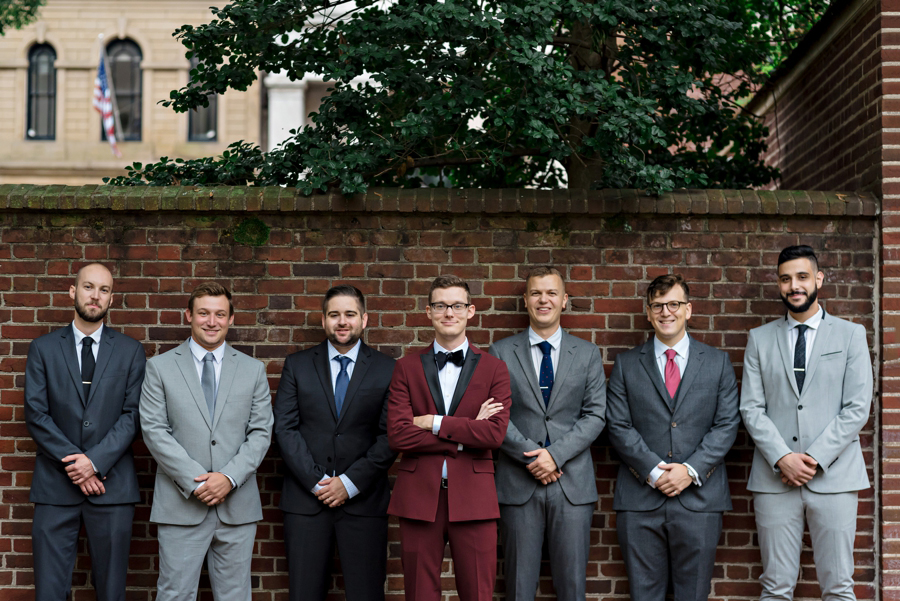 A groom in a maroon suit stands with his groomsmen agains a brick wall.